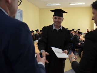 Absolwent w todze wyciąga dłoń po gratulacje podczas wręczania dyplomu.