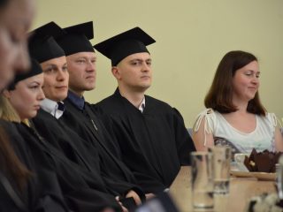 Absolwenci w czarnych togach i biretach siedzą w rzędzie przy stole.