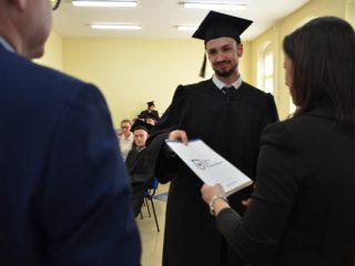 Absolwent z wąsami i brodą odbiera dyplom od organizatorki.