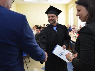 Uśmiechnięty absolwent w todze i niebieskim krawacie ściska dłoń przedstawiciela uczelni.