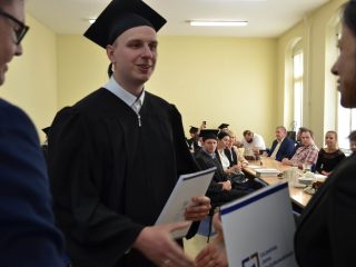 Absolwent w todze i birecie trzyma teczkę z dyplomem podczas odbierania gratulacji.