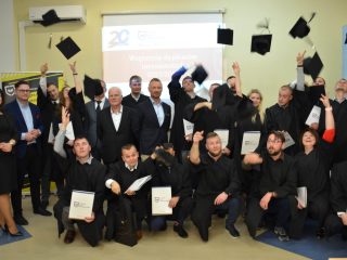 Absolwenci wyrzucają birety w górę podczas wspólnego zdjęcia z przedstawicielami uczelni.