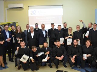 Absolwenci i przedstawiciele uczelni pozują z dyplomami; część osób klęczy w pierwszym rzędzie.