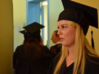 Absolwentka z długimi blond włosami, w todze i birecie, widoczna z boku w korytarzu.