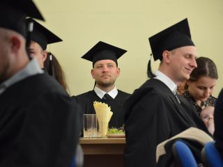 Uśmiechnięty absolwent w todze i birecie siedzi wśród uczestników uroczystości.