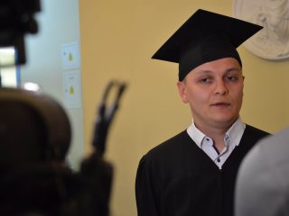 Absolwent w todze i birecie udziela wywiadu przed kamerą.