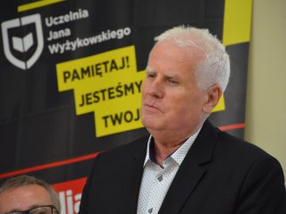 Siwowłosy mężczyzna w ciemnej marynarce na tle banera UJW.