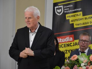Siwowłosy mężczyzna przemawia ze złożonymi dłońmi przy stole z kwiatami.