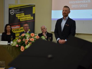 Mężczyzna w granatowej marynarce stoi przy stole prezydialnym podczas wręczenia dyplomów.