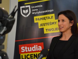 Kobieta w czarnej marynarce udziela wywiadu na tle banera UJW.