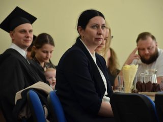Kobieta w granatowej marynarce siedzi wśród absolwentów i gości, w tym rodziny z dzieckiem.
