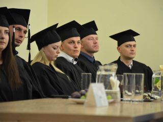 Grupa absolwentów w czarnych togach i biretach słucha wystąpienia przy stole.