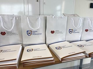 Białe torby z logo Gminy Polkowice i UJW oraz teczki z dyplomami ułożone na stole.