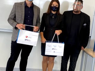 Troje uczestników uroczystości pozuje z torbami upominkowymi na tle białej tablicy.