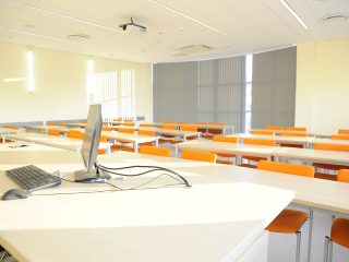 Stanowisko komputerowe prowadzącego w sali z pomarańczowymi krzesłami.
