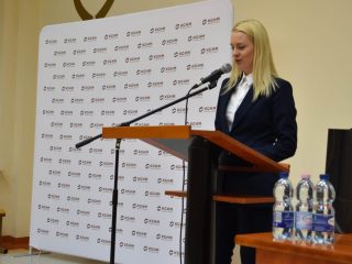 Kobieta przemawia przy mównicy obok stołu z butelkami wody w auli UJW.