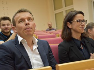 Mężczyzna w garniturze i kobieta w okularach siedzą w pierwszym rzędzie auli; kobieta klaszcze.