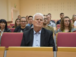 Siwowłosy mężczyzna i pozostali słuchacze wykładu siedzą w rzędach czerwonych foteli.