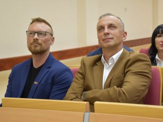 Dwaj uczestnicy wykładu w niebieskiej i brązowej marynarce siedzą obok siebie w auli.
