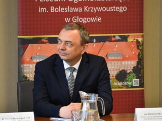 Mężczyzna siedzi przy stole na tle banera I LO w Głogowie.