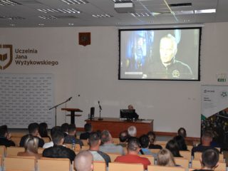 Uczestnicy wykładu oglądają film z wypowiedzią mężczyzny wyświetlany na dużym ekranie.