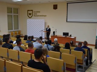 Widok auli z publicznością i kobietą przemawiającą przy mównicy.