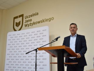 Mężczyzna w granatowej marynarce przemawia przy mównicy na tle logo UJW.