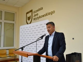 Prelegent w granatowej marynarce opiera dłonie na mównicy podczas wystąpienia.