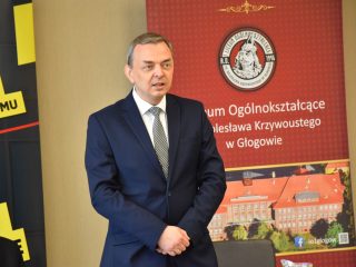 Mężczyzna przemawia na tle banera I LO im. Bolesława Krzywoustego w Głogowie.