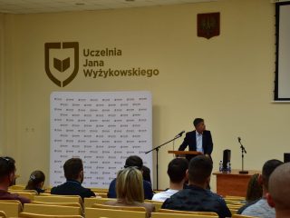 Prelegent przy mównicy spogląda w stronę ekranu podczas wykładu.