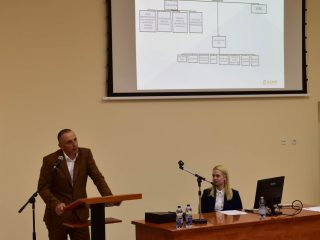 Prelegent w brązowym garniturze stoi pod ekranem ze schematem podczas wykładu o ratownictwie.
