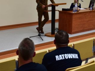 Dwaj ratownicy siedzą na widowni przed prelegentem w brązowym garniturze.