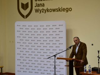 Mężczyzna w brązowym garniturze przemawia przy mównicy na tle logo UJW.