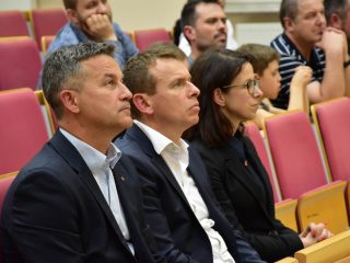 Dwaj mężczyźni w garniturach i kobieta w okularach słuchają wykładu w pierwszym rzędzie.