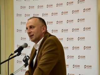 Zbliżenie na prelegenta w brązowej marynarce mówiącego do mikrofonu na tle logotypów KGHM.