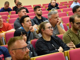 Uczestnicy wykładu siedzą w czerwonych fotelach, spoglądając w stronę prowadzącego.