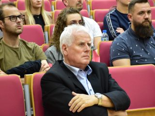 Siwowłosy mężczyzna siedzi ze skrzyżowanymi rękami wśród uczestników wykładu.