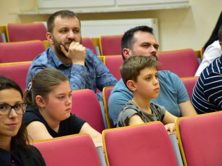 Dzieci i dorośli słuchają wykładu, siedząc w rzędach czerwonych foteli.