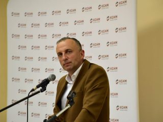 Prelegent w brązowej marynarce spogląda na słuchaczy znad mikrofonu.