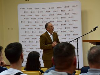 Mężczyzna w brązowym garniturze gestykuluje podczas wystąpienia przed publicznością.
