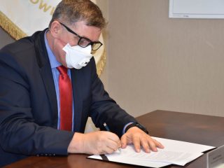 Mężczyzna w maseczce i czerwonym krawacie podpisuje dokument współpracy.