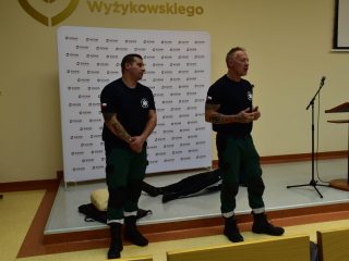 Dwaj ratownicy w czarnych koszulkach i zielonych spodniach prowadzą pokaz przed publicznością.