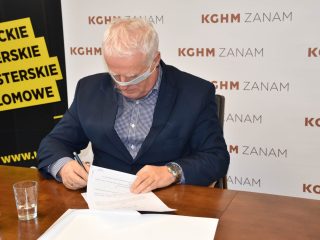 Siwowłosy mężczyzna w przyłbicy podpisuje umowę przy stole.