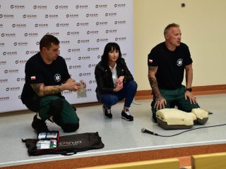 Dwaj ratownicy i uczestniczka klęczą przy fantomie oraz rozłożonym wyposażeniu do pokazu pierwszej pomocy.