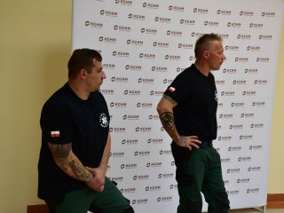 Dwaj ratownicy w czarnych koszulkach i zielonych spodniach stoją przed publicznością.