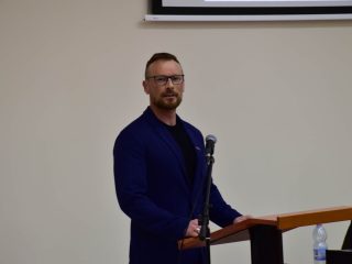 Mężczyzna w okularach i niebieskiej marynarce stoi przy mównicy z mikrofonem.