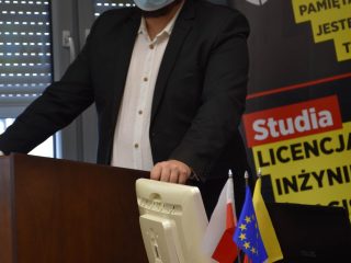 Mężczyzna w okularach prowadzi wystąpienie przy mównicy i komputerze.