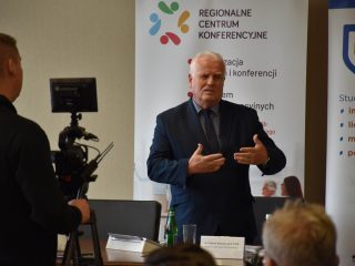 Mówca gestykuluje podczas wystąpienia przy banerze Regionalnego Centrum Konferencyjnego.