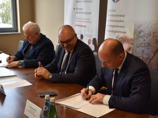 Trzej uczestnicy spotkania składają podpisy na dokumentach przy stole.
