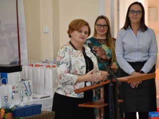Kobieta w kwiecistej marynarce przemawia do mikrofonu, a obok stoją dwie organizatorki.